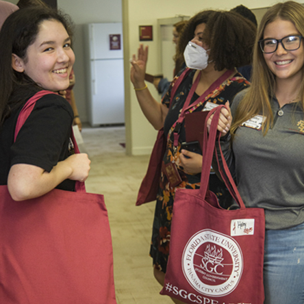 Female students smiling at the camera and standing in a building hallway holding re-useable bags
